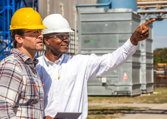 two contractors in hard hats outside looking to the right, one has an arm raised and is pointing