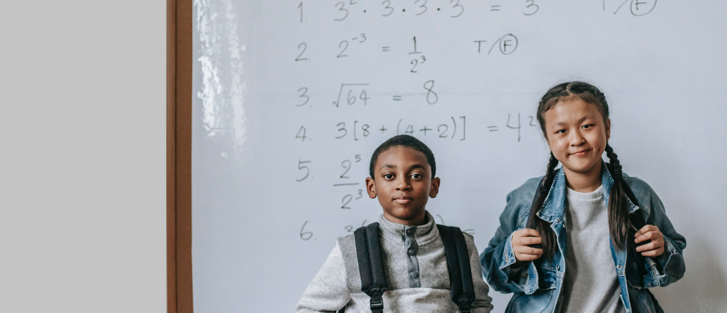 Two kids in a classroom standing in front of a whiteboard, smiling at the camera