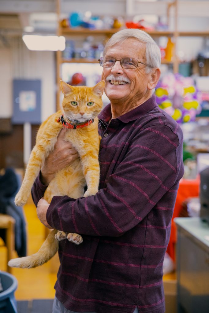 Gray-haired, smiling small business owner holding orange tabby cat in his flower shop.