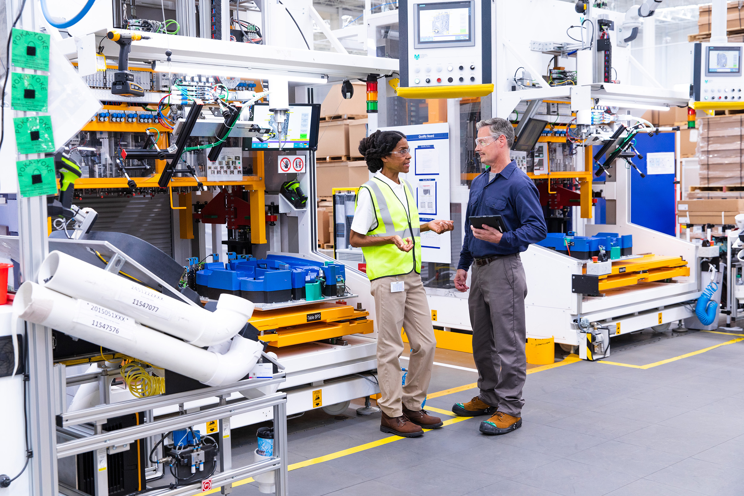Man with a tablet and woman wearing a yellow safety vest talking in an industrial facility warehouse