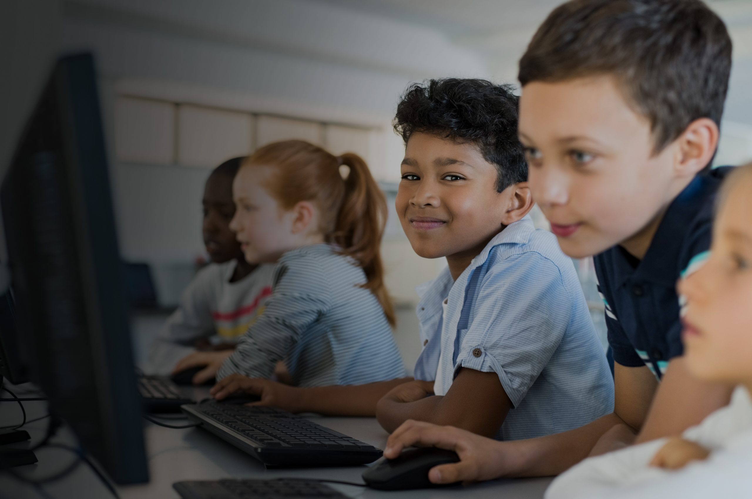 children at school, sitting in a row looking at computers, one is smiling at the camera