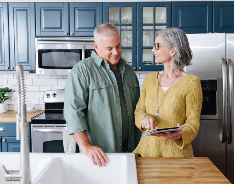 Older couple in kitchen smiling at each other, woman is pointing at tablet she is holding
