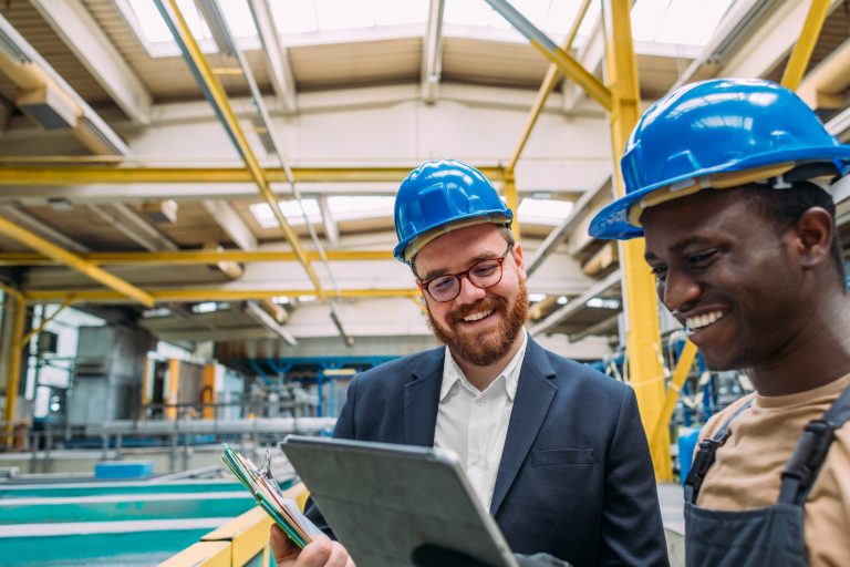 Two men stand in an industrial setting looking at a tablet