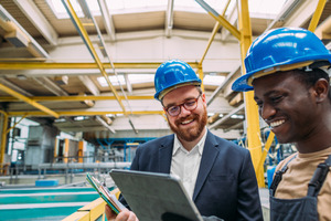 Sandy haired, bearded man with glasses and a blue hardhat in an industrial setting holding a clipboard and smiling and talking to a Black man wearing a blue hardhat and holding a tablet.