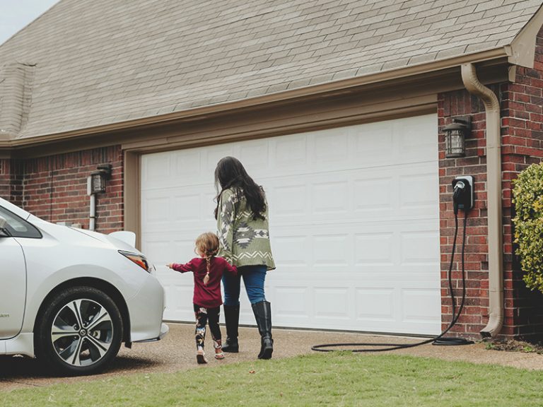 Mother and daughter walking towards their EV after unplugging it from their home charger, which is attached to the exterior of their garage.