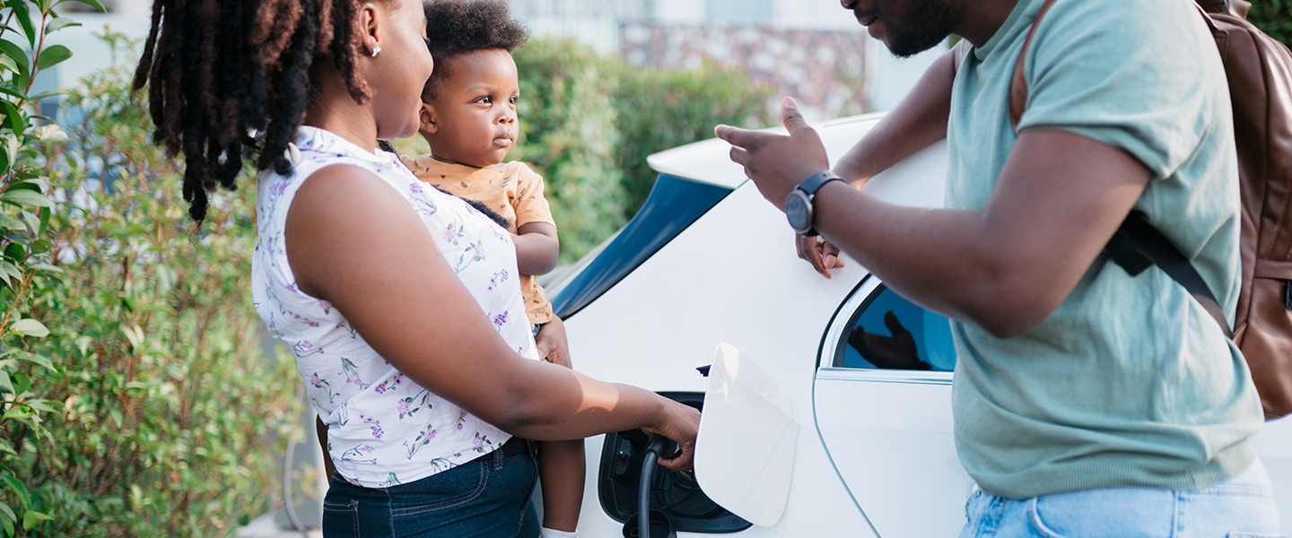 Mother holding toddler on her hip while charging the family's EV. Father is leaning on the car, chatting happily with them.