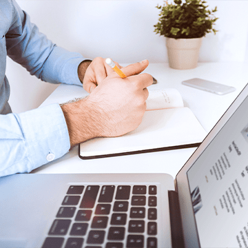 Hands writing on notebook next to open laptop