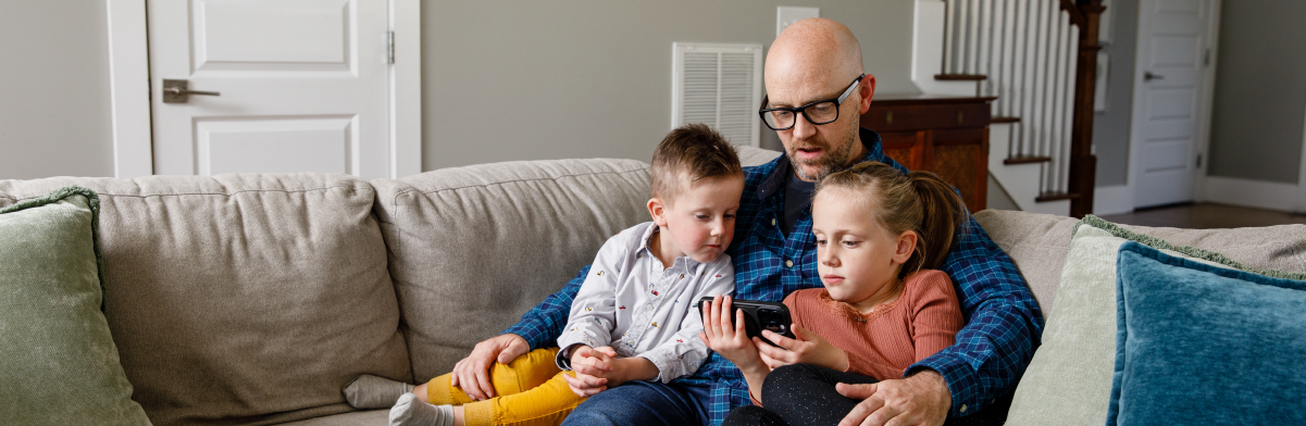 Father sitting on couch with son and daughter, all looking at cell phone screen the daughter is holding