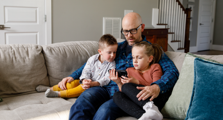 Father sitting with his son and daughter on the couch, all looking at a cell phone screen the daughter is holding