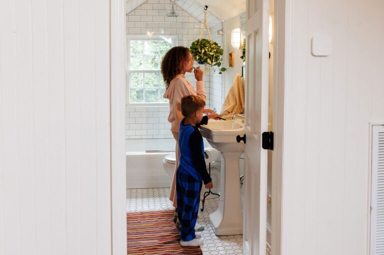 Two kids brushing their teeth in a comfortable home with a smart thermostat on the wall in the hallway outside the bathroom.