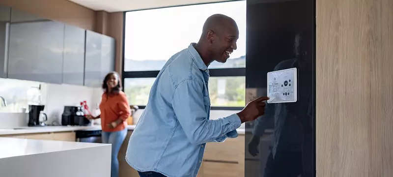 Smiling man in his bright and modern kitchen adjusting his home energy monitoring device. A woman is smiling in the background.