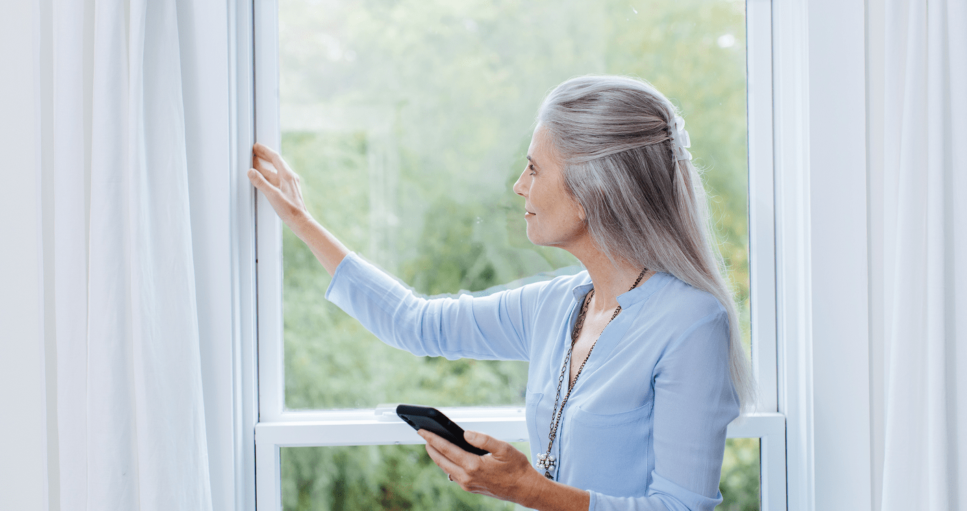 Woman with silver hair, wearing a pale blue shirt, checking for leaks along the side of her window.