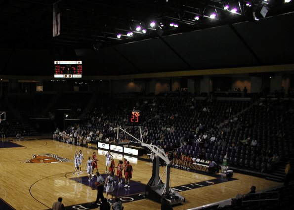 High-angle view of an indoor sports arena showing high-efficiency overhead arena lighting over a basketball game, spectator seating, and an electronic scoreboard.
