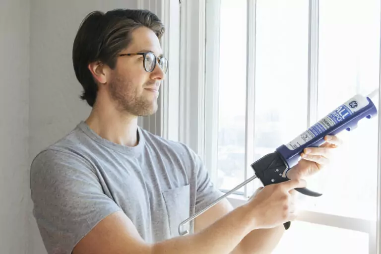 A homeowner uses a caulking gun to apply sealant along a residential window frame to reduce drafts and improve home air sealing.