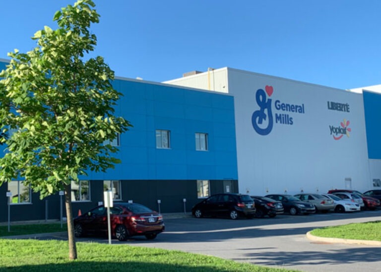 Exterior of a large General Mills industrial facility with prominent General Mills, Liberté, and Yoplait brand logos on its white and blue facade. Cars are parked in front.