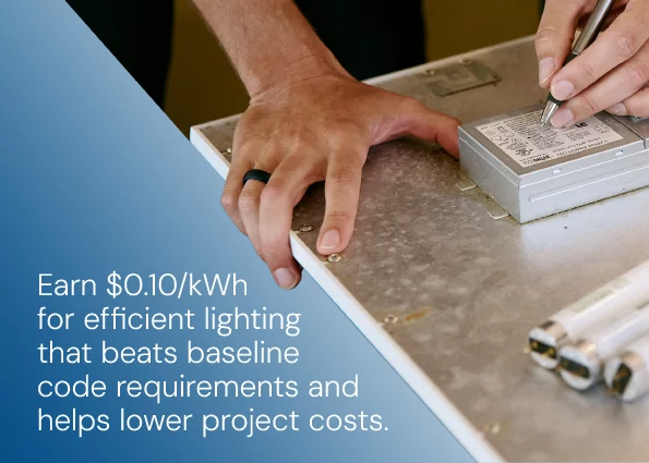 A technician uses a pen to note specifications on a lighting ballast fixture next to linear lamp tubes. Text overlay reads: Earn $0.10/kWh on installed building-wide Lighting Power Density and reduce upgrade costs.