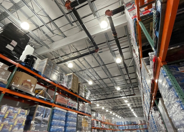 Commercial warehouse interior showing high ceilings with visible insulation, metal racking, and overhead lighting, suggesting efficient lighting upgrades.