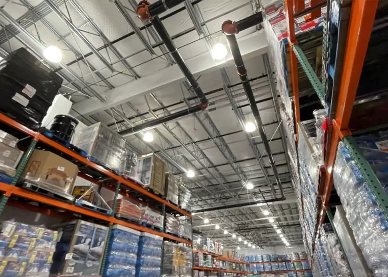 Commercial warehouse interior showing high ceilings with visible insulation, metal racking, and overhead lighting, suggesting efficient lighting upgrades.