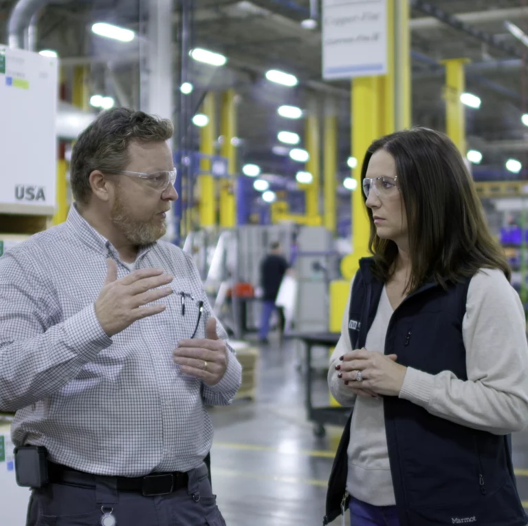 A TVA Energy Expert wearing protective eyewear gestures while discussing industrial energy-efficient upgrades with a facility representative on a factory floor.