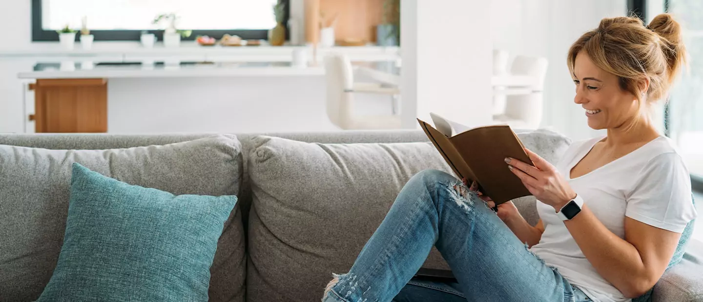 Woman smiles reading a book on a comfortable sofa, enjoying her bright, energy-efficient home environment.