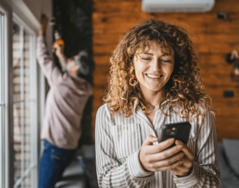 Smiling woman on phone as TVA Energy Expert installs energy-efficient window treatments near a heat pump.